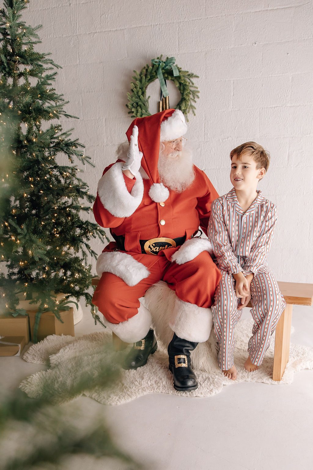 A smiling boy in pajamas sitting beside Santa in front of a Christmas tree and wreath
